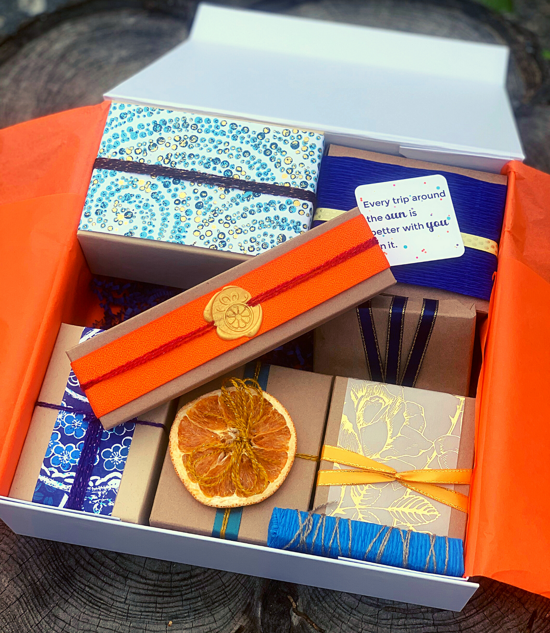 An orange hand-wrapped gift rests on top of an open box of 7 gifts